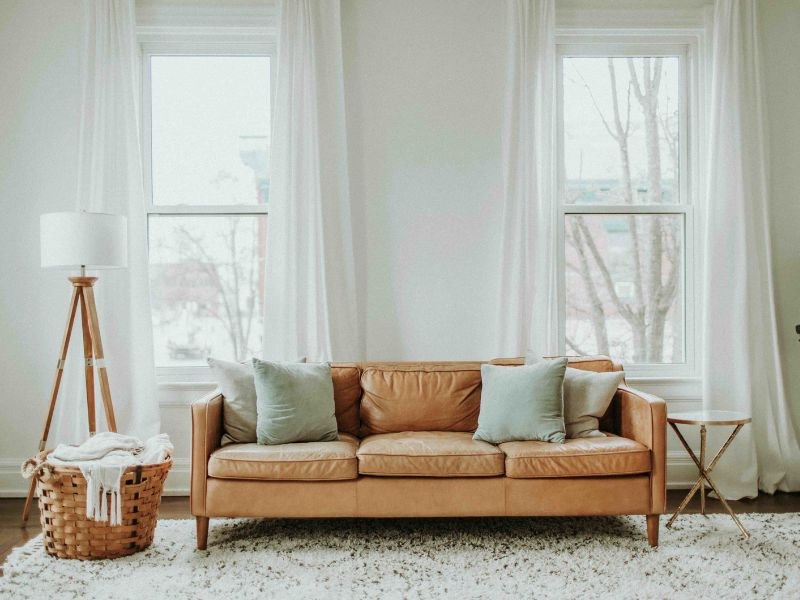 5 Dicas Definitivas de Cortina para Você Escolher a Ideal 2 5 dicas definitivas de cortinas, imagem mostra uma sala com duas janelas retangulares com cortinas brancas, transparentes. Na frente delas, um sofa de couro marrom claro , uma luminaria de piso e uma cesta de vime