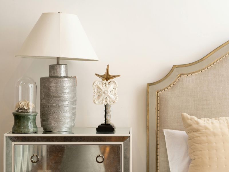 Como Arrumar Objetos na Decoração 2 como arrumar objetos, imagem mostra uma mesa de cabeceira espelhada, decorada com uma luminaria e dois objetos decorativos