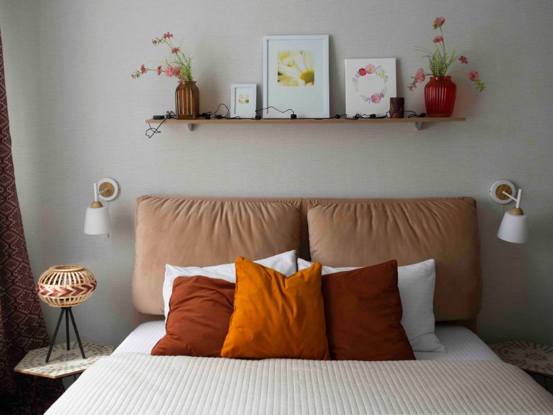 Como Montar Um Quarto De Casal: Guia Com Medidas 3 como montar um quarto de casal, imagem mostra um quarto de casal com cama, cabeceira estofada, almofadas em cores branca e terrosa, uma prateleira em cima da cama com quadros e objetos decorativos